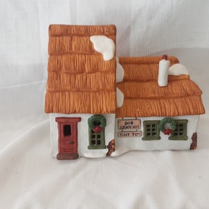 Puede incluir: Una casa de cerámica en miniatura con tejado de paja y detalles de nieve. La casa tiene una puerta roja, ventanas con marcos verdes con coronas y un letrero que dice "Bob Cratchit Tiny Tim."