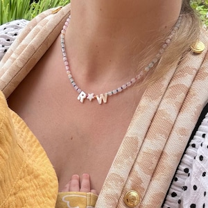 May include: A beaded necklace featuring small, multi-coloured pastel beads and three central charms made of white material. The charms consist of the letter R, a five-pointed star, and the letter W, arranged in that specific order on the strand.