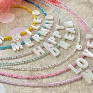 May include: Several beaded necklaces spelling out words such as "MERMAID", "HAPPY", and "LOVE" with white letter beads. The necklaces feature colourful beads in yellow, blue, green, and pink, strung on clear cords.