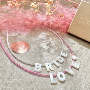 May include: A white and pink beaded necklace with the words "BRIDE" and "LOVE" spelled out in white pearl-like letters.