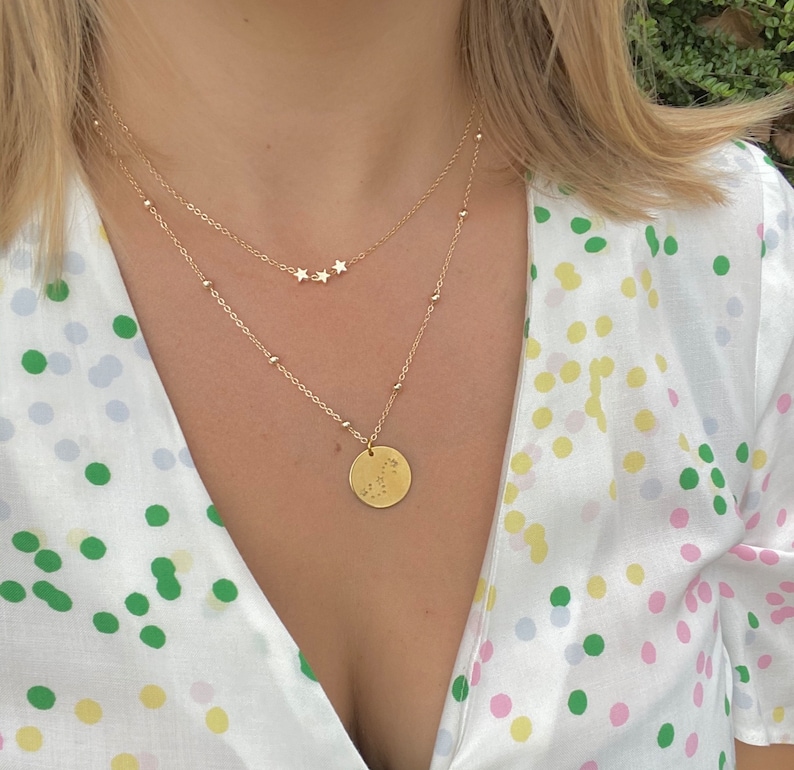 May include: A gold necklace with a small round pendant featuring a constellation design. The necklace is layered with a delicate chain with three small star charms.