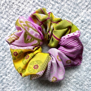 Puede incluir: Una colorida scrunchie hecha de tela estampada. La scrunchie presenta una mezcla de telas rosas, amarillas y verdes con diseños florales y geométricos. Un accesorio para el cabello.