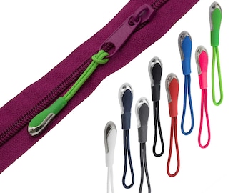 Könnte beinhalten: Sortiment an Reißverschluss-Zierbändern in verschiedenen Farben, darunter Grün, Blau, Rot, Weiß und Schwarz. Jeder Zipper hat eine silberne Metallschlaufe und eine kleine, abgerundete Plastikspitze.