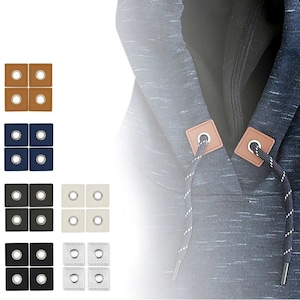 Könnte beinhalten: Satz mit vier braunen, schwarzen, weißen und blauen Leder-Kapuzenverschlüssen mit silbernen Metallösen. Die Verschlüsse sind auf einem blauen Kapuzenpullover abgebildet.