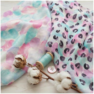 Könnte beinhalten: Zwei Stoffstücke mit Aquarellmustern in Rosa, Lila und Türkis. Ein Stoff zeigt weiche, abgerundete Formen, der andere ein dunkles Leopardenmuster. Eine Spule mit türkisfarbenem Garn, ein einzelner Knopf und Baumwollblüten sind abgebildet.