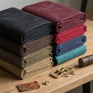 Könnte beinhalten: Ein Stapel gefalteter gewachster Canvas-Stoffe in Schwarz, Dunkelbraun, Beige, Burgunderrot, Rot, Marineblau und Petrol. Daneben liegen Messingschnallen, ein kleines Lederstück, ein Messingknopf und eine Spule mit dunkelbraunem Garn.