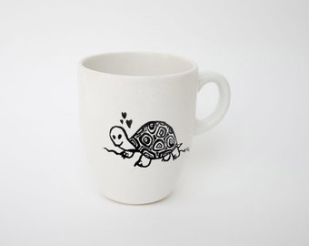 May include: White ceramic mug with a black cartoon turtle illustration. The turtle has a patterned shell and is holding a sprig with hearts above it. The mug has a curved handle and is set against a white background.