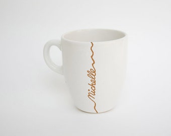 May include: White ceramic mug with a curved handle. A brown, decorative line runs vertically down the side, with the name "Michelle" in a cursive font. The mug is set against a white background.