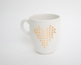 May include: White ceramic mug with a gold cross-stitch heart design. The mug has a curved handle and a smooth, glossy finish. A simple and elegant design for coffee or tea.