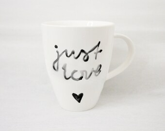 May include: White ceramic mug with the words "just love" in black script and a small black heart. The mug has a curved handle and a simple, elegant design, suitable for coffee or tea.