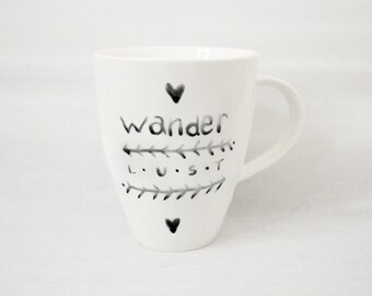 May include: White ceramic mug with a black handle. The mug features the word "wander" in a handwritten font, along with the words "L.U.S.T." and decorative elements. Two black hearts are also on the mug.