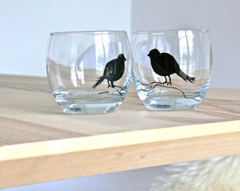 May include: Two clear stemless wine glasses with a black bird silhouette design. The birds are perched on a branch. The glasses are sitting on a light-colored wooden surface.