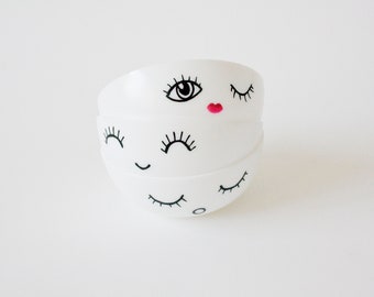 May include: Three white ceramic bowls stacked on top of each other. Each bowl is decorated with a black and white eye design, with one bowl featuring a red lip. The bowls are stacked on a white surface.