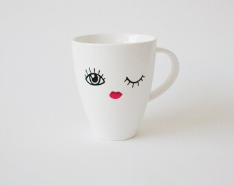 May include: White ceramic mug with a whimsical face design. The mug features a black-outlined eye, a winking eye, and red lips. The mug has a curved handle and a smooth, glossy finish.
