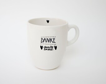 May include: White ceramic mug with a handle, featuring black text that reads "DANKE dass Du DA bist." and small heart illustrations. A single heart is near the rim. The mug is designed for beverages.
