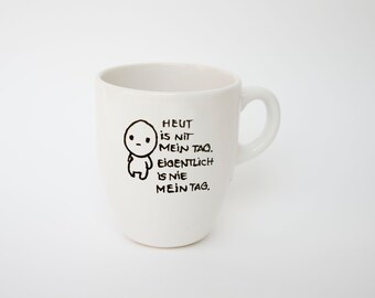 May include: White ceramic mug with a handle, featuring a black cartoon figure and text that reads "Heut is nit mein Tag. Eigentlich is nie mein Tag."