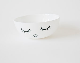 May include: A white ceramic bowl with a whimsical design. The bowl features a simple face with closed eyes and a small circle for a nose, all drawn in black. The bowl has a smooth, glossy finish.