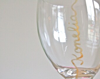 May include: Clear wine glass with the name "Konelia" written in gold script. The glass is partially filled and reflects the surrounding environment. The stem and base are visible.