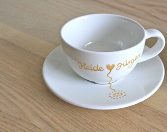 May include: White ceramic teacup and saucer set. The cup and saucer have gold lettering and a heart design. The text on the cup reads "Heide Jürgen" and the saucer has a "50" sunburst design.