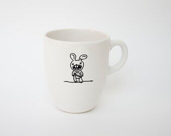 May include: White ceramic mug with a black cartoon bunny illustration. The bunny has a stitched mouth and cross eyes. The mug has a curved handle and a smooth, glossy finish. The design is centered on the mug.