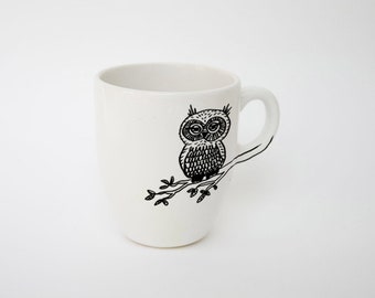 May include: White ceramic mug with a black illustration of an owl perched on a branch. The owl has detailed feathers and large eyes. The mug has a curved handle and a simple, clean design.