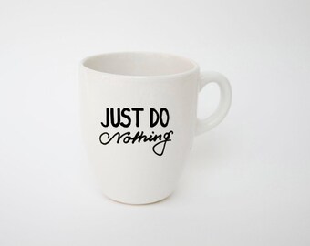 May include: White ceramic mug with a curved handle. The mug features the black text "JUST DO Nothing" in a handwritten style. The mug is set against a plain white background.
