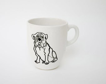 May include: White ceramic mug featuring a black line drawing of a bulldog. The dog is sitting with its front paws forward. The mug has a curved handle and a cylindrical shape, suitable for coffee or tea.