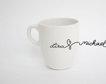 May include: White ceramic mug with a curved handle. The mug features the names "Lisa" and "Michael" in a continuous black script, with a heart symbol connecting the names. The mug is set against a plain white background.