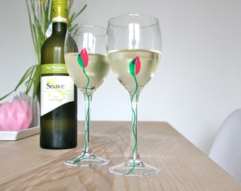 May include: Two wine glasses filled with white wine, each adorned with a hand-painted red and green floral design. A bottle of white wine and a pink candle are in the background, set on a light wood table.