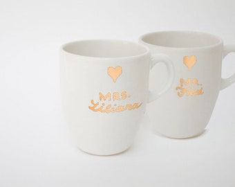 May include: Two white ceramic mugs with gold lettering and heart accents. One mug reads "MRS. Luciana", the other reads "MR. Fred". The mugs have a simple, elegant design, perfect for a wedding or anniversary gift.