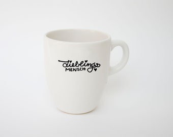 May include: A white ceramic mug with a curved handle. The mug features the black text "Lieblingsmensch" with a heart symbol. The mug is set against a plain white background, highlighting its simple design.