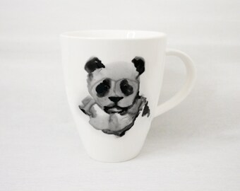 May include: White ceramic mug featuring a black and gray watercolor illustration of a panda wearing glasses. The panda design is centered on the mug. The mug has a curved handle and a smooth, glossy finish.