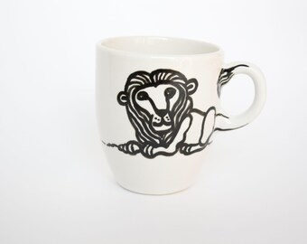 May include: White ceramic mug with a black lion illustration. The lion is depicted in a relaxed pose, with detailed linework. The mug has a curved handle and a simple, clean design, suitable for coffee or tea.