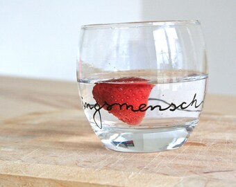 May include: A clear glass filled with water, with a vibrant red strawberry submerged inside. The glass has the word "ingsmensch" written on it in black script. The glass sits on a light brown wooden surface.