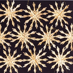 Könnte beinhalten: Sieben Stroh-Schneeflocken mit goldenen Fadenschlaufen sind auf einem schwarzen Hintergrund angeordnet. Jede Schneeflocke hat ein Sternmuster, wobei mehrere Strohhalme von einem zentralen Punkt ausgehen. Die Schneeflocken sind hellbraun.