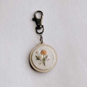 Może przedstawiać: Mały, okrągły drewniany brelok z białym kołem z tkaniny w środku. Koło z tkaniny ma haftowany wzór kwiatowy z pomarańczową, białą i zieloną nicią.