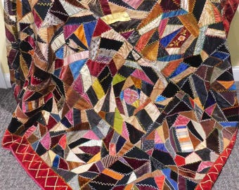 Pode incluir: Um edredão de retalhos colorido com uma variedade de formas geométricas em tons de castanho, preto, vermelho e azul. O edredão tem uma borda vermelha com um desenho de ponto cruz dourado. O edredão é feito de veludo e outros tecidos.