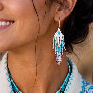 Op de afbeelding: Een meerrijige kralenketting en een bijpassende oorbel in kroonluchterstijl, bestaande uit witte, turquoise, lichtpaarse en metallic bronzen kralen. De sieradenset is gemaakt van kleine rocailles die in een gelaagd patroon zijn gerangschikt.