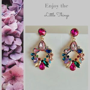 Könnte beinhalten: Ein Paar goldfarbene Ohrhänger mit einer kreisförmigen Anordnung aus facettierten, bunten Kristallen in Rosa, Blau, Grün und klaren Tönen. Der Text "Enjoy the Little Things" ist darüber auf einem hellgrauen Hintergrund aufgedruckt.