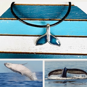 Op de afbeelding: Een zilveren walstaart-hangerketting met een blauwe emaille afwerking. De ketting is aan een zwart leren koord bevestigd en wordt getoond op een blauw-wit gestreepte achtergrond. De afbeelding toont ook twee walstaarten in het water.