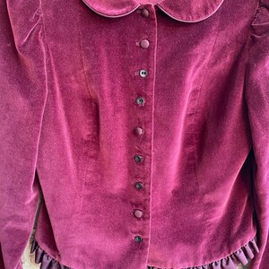 May include: A burgundy velvet jacket with a peter pan collar and a row of black buttons down the front. The jacket has a ruffled hem.
