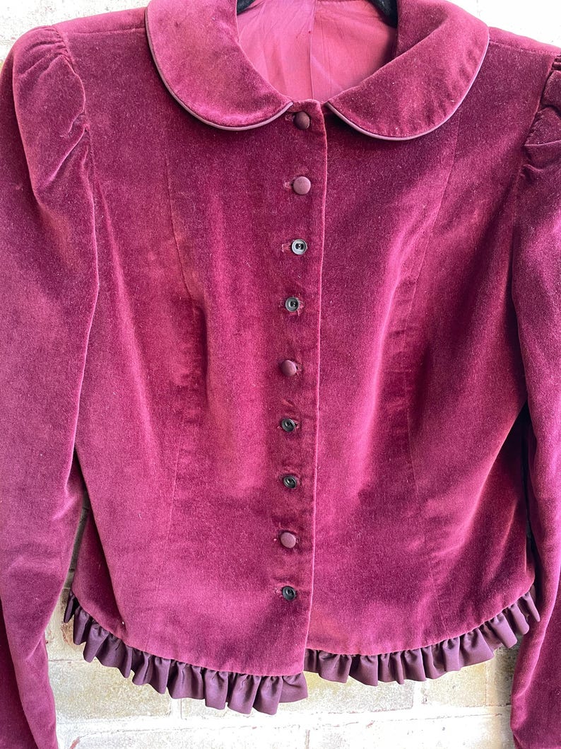 May include: A burgundy velvet jacket with a peter pan collar and a row of black buttons down the front. The jacket has a ruffled hem.