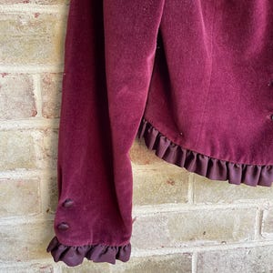 May include: A burgundy velvet jacket with a ruffled trim around the bottom and sleeves. The jacket has a button closure on the sleeve.