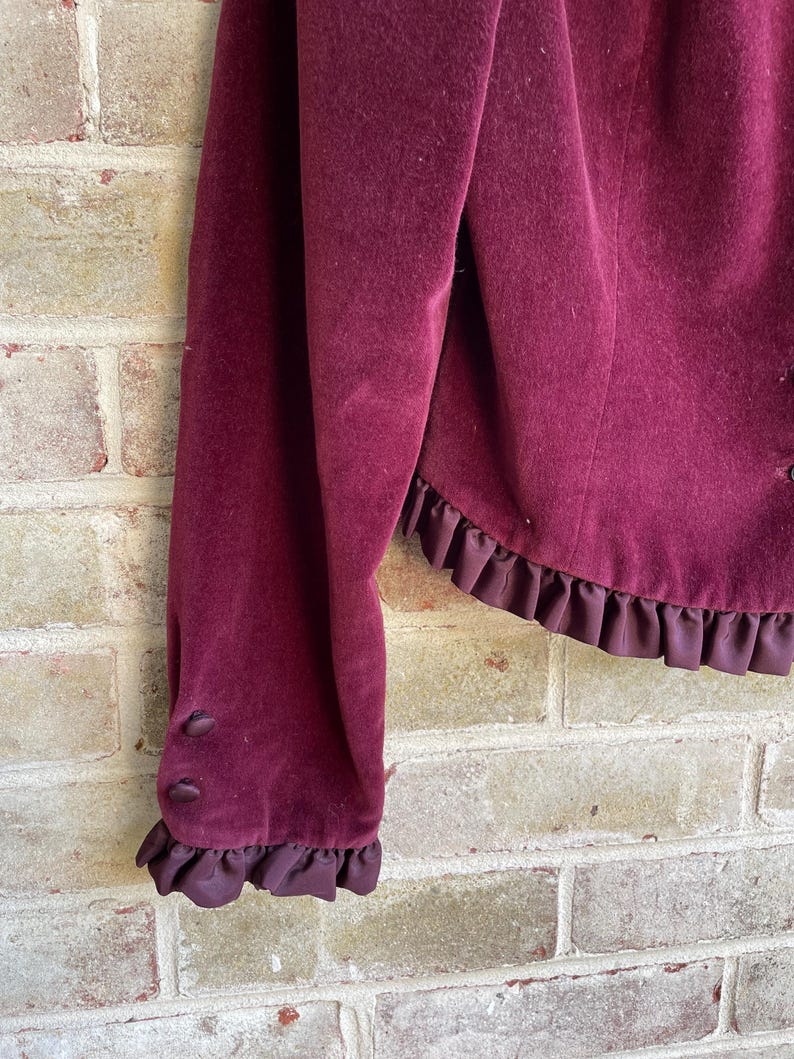 May include: A burgundy velvet jacket with a ruffled trim around the bottom and sleeves. The jacket has a button closure on the sleeve.