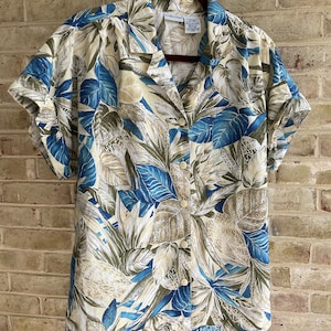 May include: A short-sleeved, button-up shirt with a tropical print featuring blue and green leaves and a beige background. The shirt has a collar and a button closure. The label reads "CARIBBEAN CLUB".