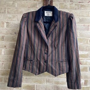 May include: A long-sleeve blazer featuring a vertical striped pattern in shades of brown, black, and red. The garment has a solid dark navy blue collar, three front buttons, and a pointed hemline. A white label inside reads Kasper ASL Petite.