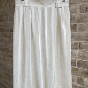 May include: A cream-colored, vertically striped skirt hanging on a hanger. The skirt features a tailored waistband with belt loops and a pleated design. The fabric has a textured appearance, with alternating cream and off-white stripes.