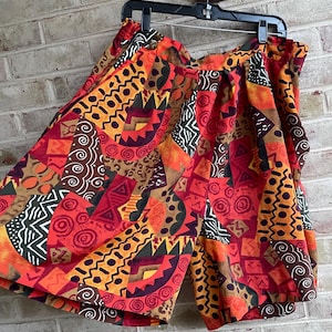 May include: A pair of colorful shorts with an abstract geometric pattern in shades of red, orange, brown, and yellow. The shorts are hanging on a black hanger against a brick wall background.