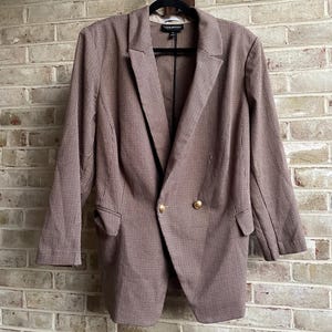 May include: A brown and white plaid blazer with gold buttons. The double-breasted jacket is hanging on a black hanger against a brick wall. The blazer has two front pockets and a notched lapel.