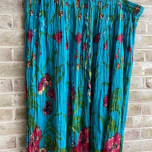 May include: A turquoise skirt with a floral pattern of pink and yellow flowers and green leaves. The skirt is hanging on a hanger against a brick wall. The skirt has a drawstring waist.